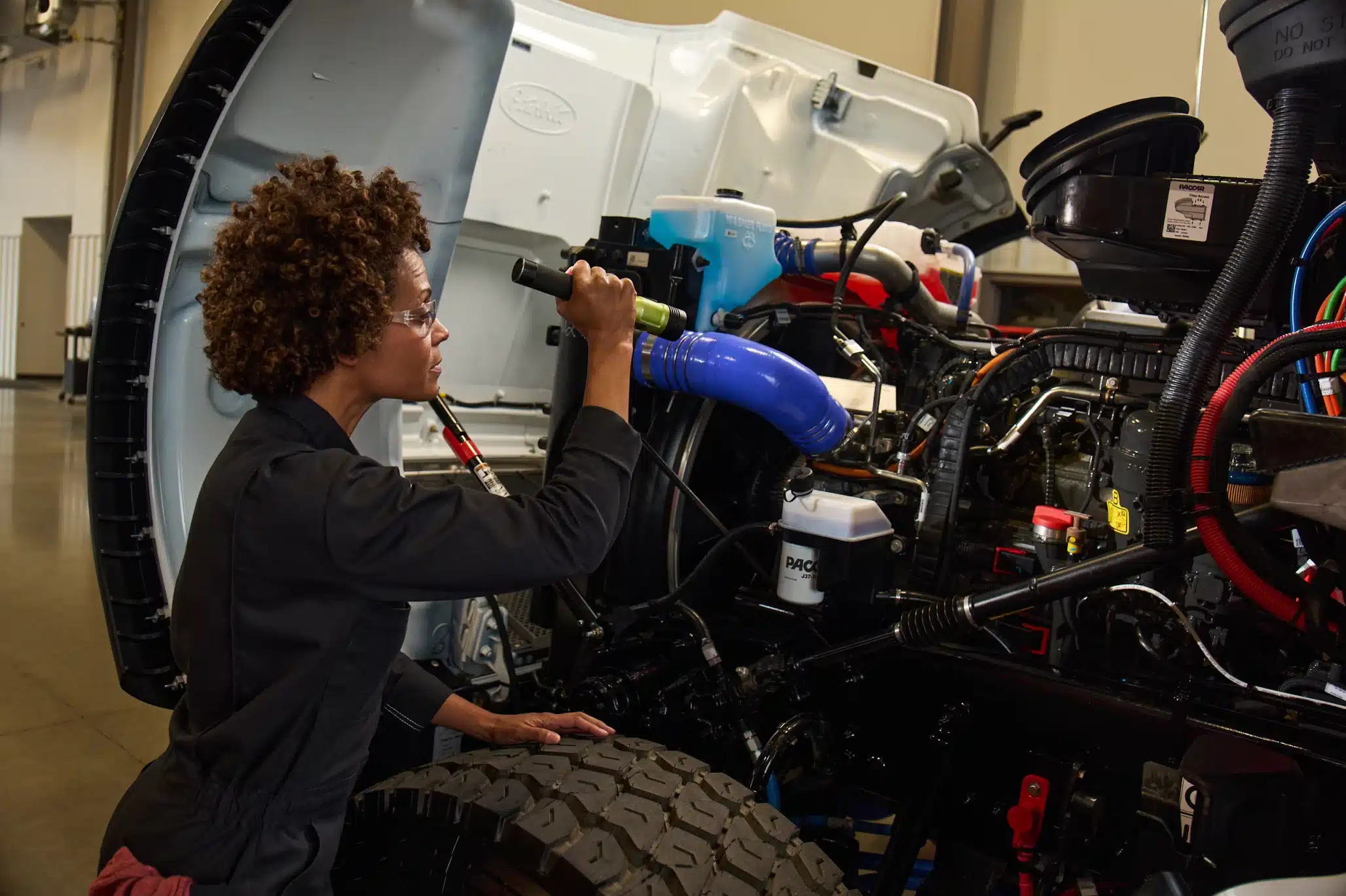 Technician inspecting a semi-truck engine compartment with a flashlight during a maintenance and diagnostic service check.