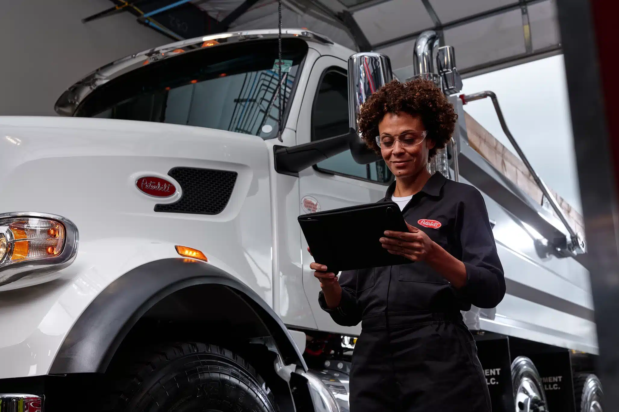 Commercial truck technician reviewing service information on a tablet while standing beside a white Peterbilt truck in a maintenance facility.