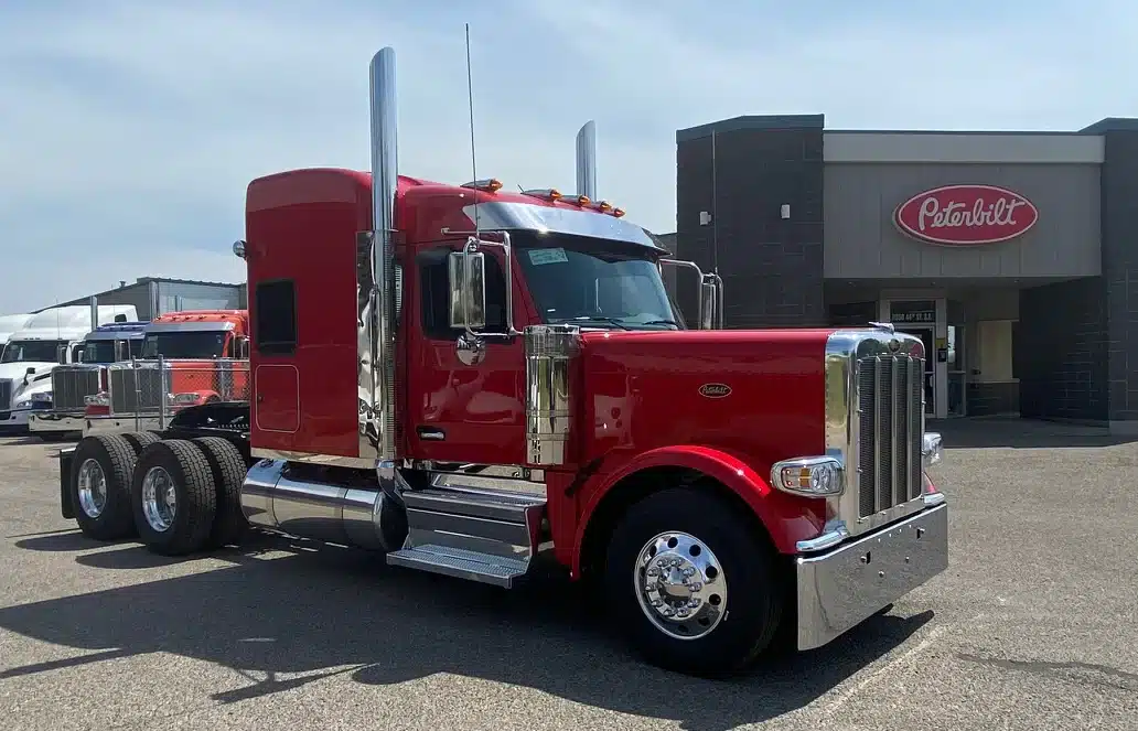 Red Peterbilt semi-truck tractor parked outside a Peterbilt dealership, showcasing a long-nose design with chrome accents and dual exhaust stacks.